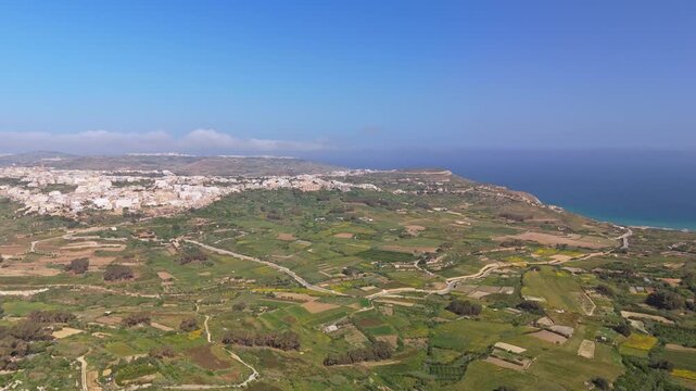 In Nadur on Gozo, Malta, terraced fields and rural buildings descend toward Ramla Bay along the Mediterranean coast, seen from an aerial perspective under clear summer skies.