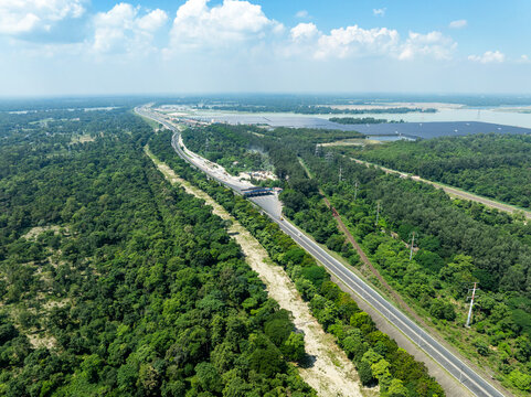 Aerial view of a long highway and toll plaza surrounded by dense green forest and lush vegetation under a blue sky with white clouds in Tangail District, Dhaka Division, Bangladesh.