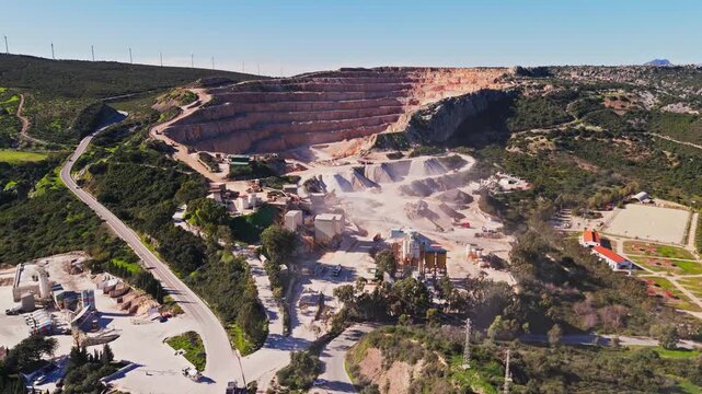 Aerial view of a large open-pit stone quarry with terraced walls, industrial processing facilities, and an asphalt plant surrounded by green hills in Manilva, Andalusia, Spain.