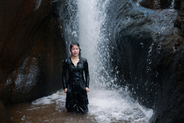 Young woman wearing black long sleeve shirt standing alone calm peaceful mood beside flowing waterfall cascade splashing down between dark rocky canyon walls beautiful natural outdoor environment © CoreRock