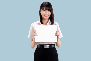 Fototapeta premium Smiling young Asian woman in student uniform holding digital tablet with blank white screen for mockup. Happy female university student showing tablet computer isolated on blue background.
