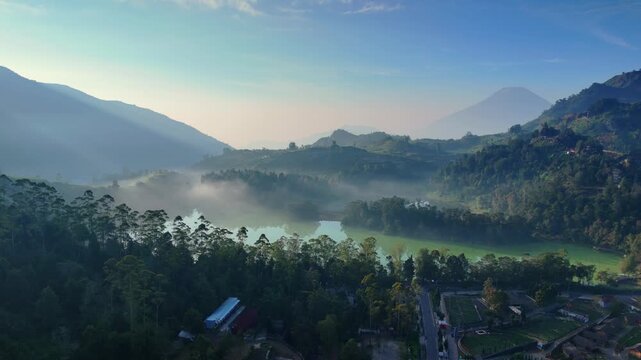 Aerial view of a serene lake surrounded by misty hills, lush forests, and distant mountains under a clear morning sky in Pekalongan, Central Java, Indonesia.