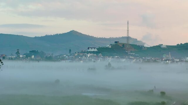 Aerial view of a village nestled on a hillside shrouded in thick morning mist with a telecommunications tower and distant mountains in Pekalongan, Central Java, Indonesia.