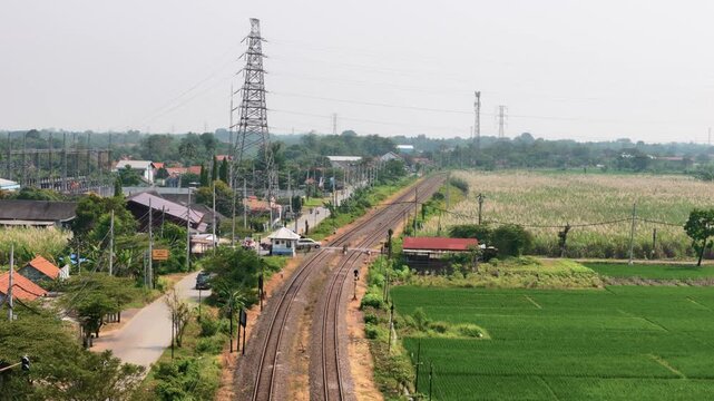 Aerial view of railway tracks passing through green fields and residential areas with a high voltage power tower in Pekalongan, Central Java, Indonesia.