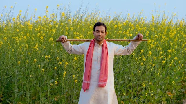 Portrait of a confident smiling farmer standing with a stick in his mustard field. Medium shot - Indian villager happily posing while holding the wooden stick on his shoulders in the Sarso field