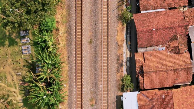 Aerial view of railway tracks passing between a cemetery and residential houses with red rooftops in Pekalongan, Central Java, Indonesia.