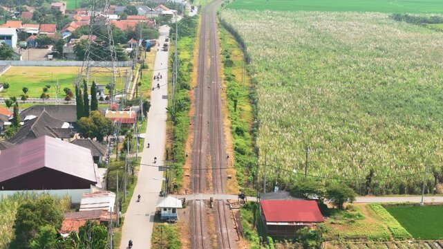 Aerial view of railway tracks running through rural landscape with houses, motorbikes on a parallel road, and lush green fields in Pekalongan, Central Java, Indonesia.