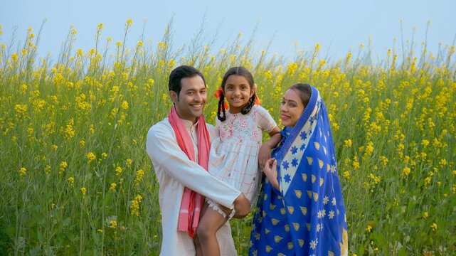 A happy rural couple lovingly holding their little daughter in their arms. Medium shot of an Indian village family happily standing in front of mustard (Sarso) agricultural field - village scene