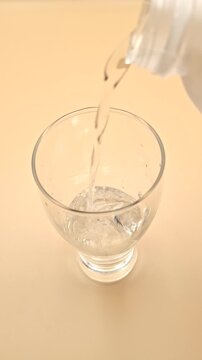 High angle shot of pouring fresh water from bottle into glass on cream background, vertical 4K, 17 seconds.