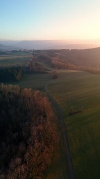 Aerial view of a winding road through green fields and dense forests during a golden sunset in Hechingen, Baden-Wurttemberg, Germany.