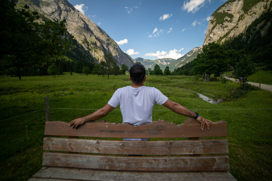 View of a man sitting on a wooden bench looking at the lush green meadows and high mountain peaks of the Eng Alm valley in Eng, Tyrol, Austria.