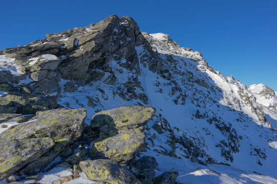View of Slavkovsky nos peak with snow-covered granite rocks and lichen-covered boulders under a clear blue winter sky in Vysoke Tatry, Presov Region, Slovakia.