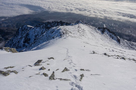 View of the snow-covered ridge of Slavkovsky nos with footprints leading toward rocky peaks above a sea of clouds in Vysoke Tatry, Presov Region, Slovakia.