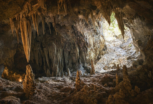 Limestone cave interior filled with stalactites and stalagmites under natural light filtering from the entrance into the rocky cavern space