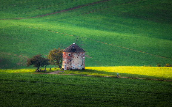 View of the historic Kunkovice windmill standing in rolling green fields with a person walking on a path under soft golden light Kunkovice, Zlin Region, Czechia.
