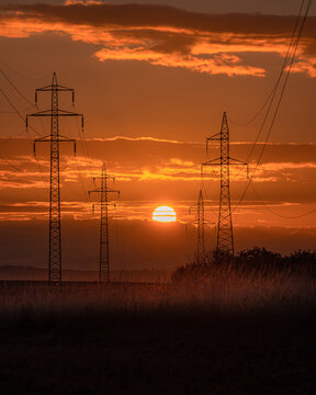 View of high voltage power lines and electricity pylons silhouetted against a vibrant orange sunset sky with clouds over a rural field in Kyjov, South Moravian Region, Czechia.