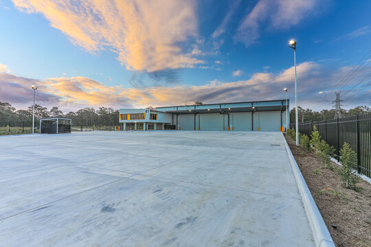 Wide-angle view of a modern logistics warehouse facility with extensive vehicle access, hardstand areas and contemporary industrial architecture beneath a dramatic sunset sky.