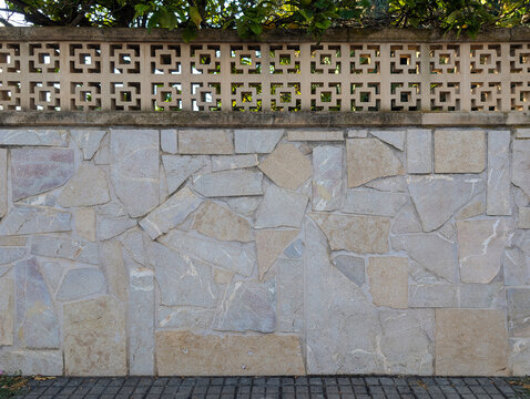 Mediterranean villa perimeter wall with natural stone cladding and geometric concrete lattice fence. Luxury architecture, landscaping, exterior construction, decorative pattern, residential enclosure.