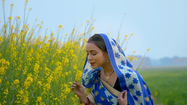 Village female farmer touching and smelling her fully grown mustard flowers. Young rural housewife in a traditional saree  happy to see her crops bloom - Indian farming concept