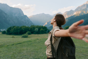 Young woman with backpack stands in a sunlit meadow with distant mountains, arms outstretched as if embracing a moment of carefree outdoor freedom. © SHOTPRIME STUDIO