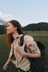 Naklejka premium A young woman with a backpack hikes through a grassy meadow with distant mountains, smiling at the peaceful landscape during golden hour.