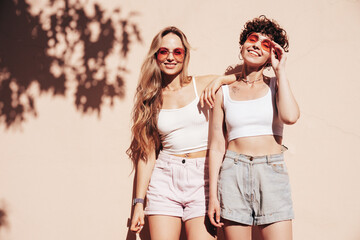 Two young beautiful smiling hipster female in trendy summer white t-shirt and shorts clothes. Sexy carefree women posing in the street. Positive models having fun, hugging and going crazy, near wall © halayalex