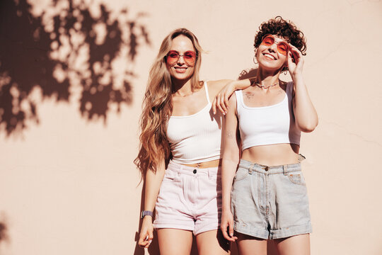 Two young beautiful smiling hipster female in trendy summer white t-shirt and shorts clothes. Sexy carefree women posing in the street. Positive models having fun, hugging and going crazy, near wall