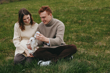 Naklejka premium Lifestyle scene with a couple feeding a goat kid outdoors on green grass, sharing affection and smiles in a bright daytime setting, portraying warm family moments outside.
