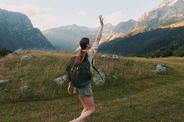 Fototapeta premium Young woman hiking outdoors in mountains, wearing backpack and casual clothes, raising hand, enjoying nature, sunny day, green grass and rocky peaks background