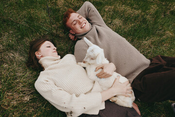 Naklejka premium Couple lies on grass enjoying a cozy moment outdoors with a friendly goat kid between them, conveying simple lifestyle intimacy amid a natural rural setting and calm countryside mood.