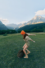 Obraz premium Young woman with long hair wearing an orange beanie jumps playfully on a backpack in a green meadow surrounded by mountains under a clear sky.