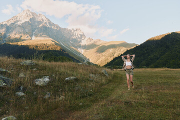 Obraz premium Young woman stands in a grassy meadow with towering mountains in the background, looking relaxed and confident.