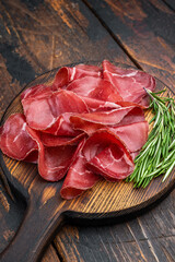 Fototapeta premium Thin cured beef slices bresaola meat arranged on a rustic wooden board with fresh rosemary over a dark textured background.