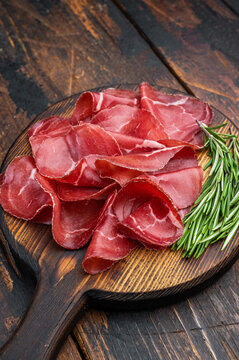 Thin cured beef slices bresaola meat arranged on a rustic wooden board with fresh rosemary over a dark textured background.