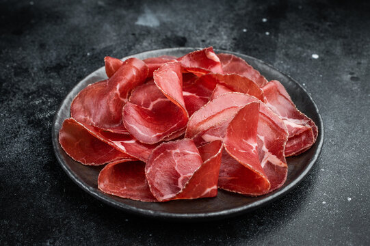 Gourmet bresaola cured beef on a dark plate over a concrete background, highlighting marbling and artisanal quality.