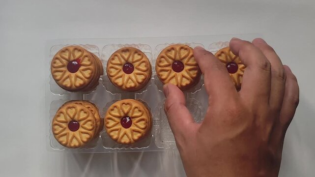 Top down view of a hand arranging and filling empty slots in a clear plastic packaging tray with jam-filled cookies on a white background.