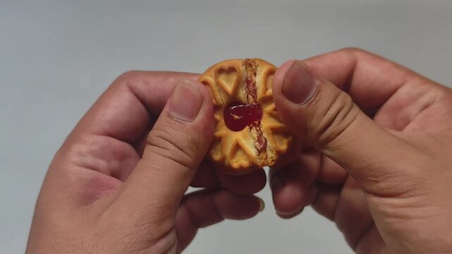 Macro close-up of hands breaking a baked butter cookie revealing sticky red jam filling inside on a clean white background, satisfying commercial food concept