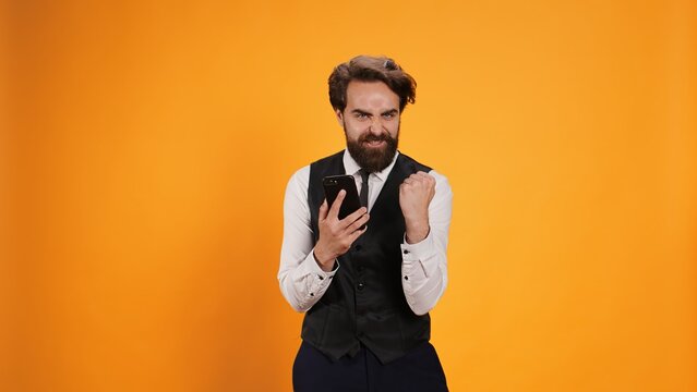 Happy waiter reads news on phone and feeling pleased on camera, preparing to serve consumers. Modern elegant butler with tie using smartphone to stay connected, social media app.
