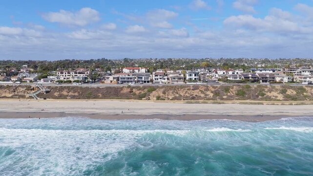 Scenic flight above Carlsbad California showing blue ocean waves breaking on sandy beach and coastal houses lining bluffs along Pacific Ocean seashore