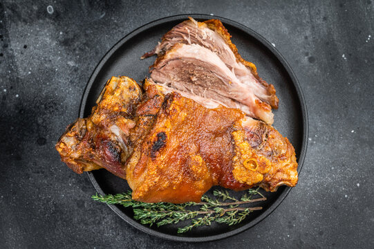 Crispy Schweinshaxe showing blistered skin, rendered fatcap and tender sliced interior, plated on dark textured background with herbs.