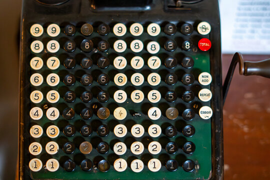 Close-up shot of a vintage mechanical cash register keyboard with vertical rows of black and white numbered keys, green background, red total button and side crank handle.