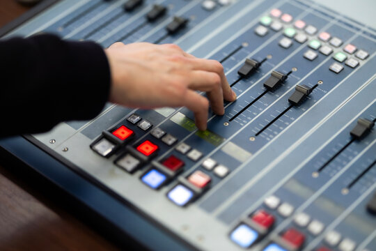 A close-up image captures an individual's hand in motion, adjusting various fader controls and engaging illuminated buttons on a professional sound mixing console used for audio production tasks