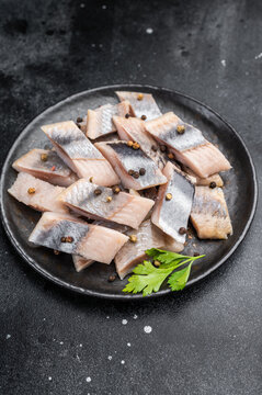 Pickled herring chunks cured in salt and brine served with spices and herbs. Traditional seafood on dark textured background.