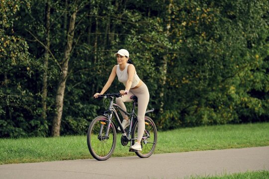 A slender woman is riding her bicycle on a smooth pathway bordered by lush greenery. She is dressed in athletic wear, enjoying the calm atmosphere of the park on a sunny day