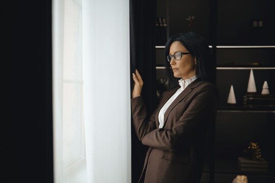 A professional woman dressed in a brown suit stands thoughtfully by a window, gazing outside