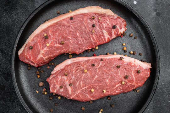Raw picanha beef steaks also known as top sirloin cap and rump cap with rosemary and pepper on black plate, dark stone background
