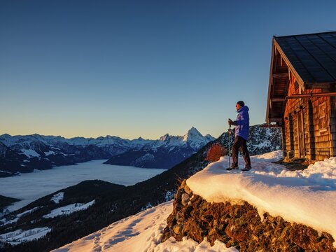 Mountaineers enjoy the sunrise in front of an alpine hut in winter, the Watzmann behind, fog in the valley, Berchtesgaden Alps, Scheibenkaser, Marktschellenberg, Berchtesgadener Land, Upper Bavaria, Bavaria, Germany
