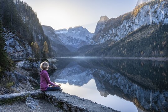 A woman doing yoga by the lake. Lotus position (Padmasana) . Vorderer Gosausee in autumn with a view of the Dachstein mountains. The Gosaukamm on the right. Blue sky, good weather. Reflection. Morning atmosphere. Vorderer Gosausee, Gosau, Gosau valley, Salzkammergut, Upper Austria, Austria