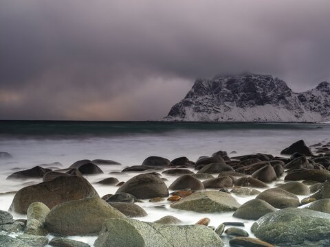 Rounded rocks on Utakleiv beach in a dramatic cloudy atmosphere, snow-capped mountains in the background, Vestvagoy Island, Lofoten, Norway