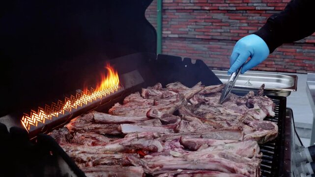 Professional chef wearing blue gloves grilling lamb ribs on outdoor barbecue, turning meat with tongs over sizzling flames, demonstrating precise culinary technique